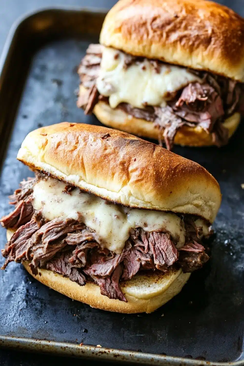 Hot roast beef sandwich slow cooker - the image shows two sandwiches on a black baking tray. the sandwiches are made with a toasted bun and are filled with shredded beef and melted cheese. the beef appears to be cooked medium-rare and the cheese is a creamy white color. the tray is lined with parchment paper and there is a blurred background.
