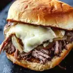 Hot roast beef sandwich slow cooker - the image shows a close-up of a sandwich on a black slate plate. the sandwich is made with a toasted bun and is topped with shredded beef, sliced onions, and melted cheese. the beef appears to be cooked medium-rare and the onions are caramelized. the cheese is melted and oozing out of the sides of the sandwich. the plate is sitting on a wooden table with a blurred background.