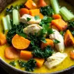 Immune boosting chicken tumeric soup - the image is a close-up of a bowl of soup. the soup appears to be a creamy yellow color with chunks of chicken and vegetables mixed in. the vegetables include carrots, celery, and kale. the bowl is made of dark brown ceramic and has a textured surface. the background is dark, making the colors of the soup stand out.