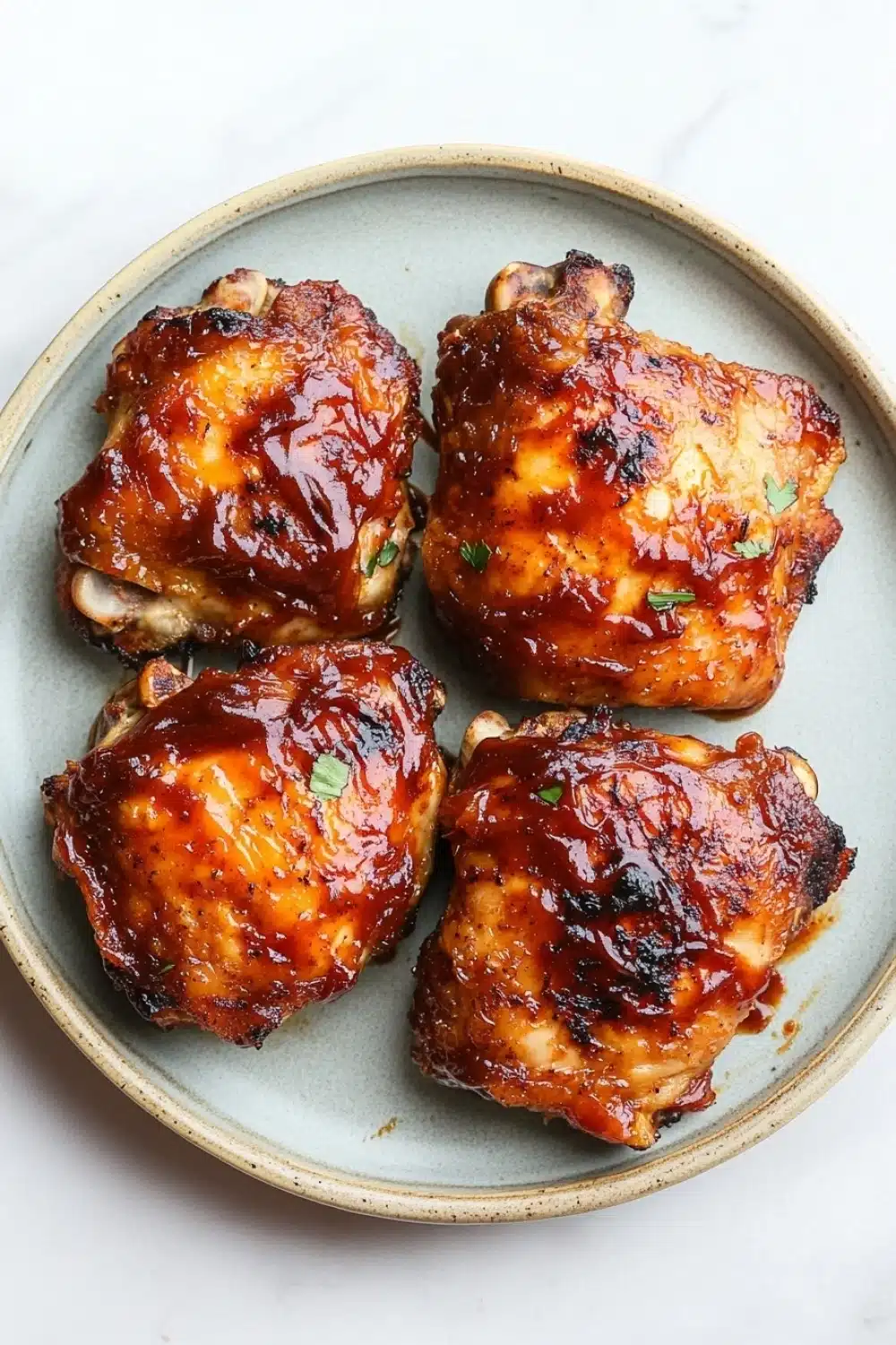 Keto baked bbq chicken thighs - the image shows four pieces of cooked chicken drumsticks on a light blue plate. the drumsticks are golden brown and appear to be seasoned with herbs and spices. they are arranged in a circular pattern on the plate, with each piece slightly overlapping the one below it. the plate is sitting on a white marble countertop.
