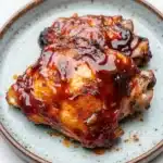 Keto baked bbq chicken thighs - the image shows two pieces of cooked chicken on a blue and white plate. the chicken appears to be seasoned with herbs and spices, and is covered in a thick, red sauce. the sauce is drizzled over the chicken, giving it a glossy sheen. the plate is sitting on a white marble countertop.