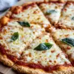 Keto pizza crust gluten free - the image shows a freshly baked pizza on a wooden cutting board. the pizza has a golden brown crust and is topped with melted cheese and fresh basil leaves. the cheese is melted and bubbly, and the basil leaves are arranged in a circular pattern around the edges of the pizza. the background is blurred, but it appears to be a kitchen countertop with a white cloth napkin on the side.