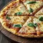 Keto pizza crust gluten free - the image shows a freshly baked pizza on a wooden cutting board. the pizza has a golden brown crust and is topped with melted cheese and fresh basil leaves. the cheese is melted and bubbly, and the basil leaves are scattered around the edges of the pizza. the cutting board is placed on a dark wooden table, and there are a few more basil leaves scattered around it. the overall mood of the image is rustic and appetizing.