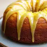 Lemon cake with lemon drizzle - the image is of a bundt cake on a blue plate. the cake is round and has a golden brown color. it is drizzled with a yellow glaze, which is dripping down the sides of the cake in a zigzag pattern. the glaze appears to be made of lemon zest, giving the cake a glossy and shiny appearance. the plate is sitting on a wooden table, and the background is blurred, making the cake the focal point of the image.