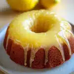 Lemon cake with lemon drizzle - the image shows a round cake on a white plate. the cake appears to be a bundt cake with a golden brown crust and a glaze on top. the glaze is dripping down the sides of the cake, creating a smooth and glossy texture. in the background, there are three whole lemons on a wooden table. the overall color scheme of the image is warm and inviting.
