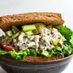 Loaded chicken salad sandwich - the image shows a sandwich in a black ceramic bowl. the sandwich is made with two slices of toasted bread and is filled with a mixture of lettuce, tomato, cucumber, and other vegetables. the salad appears to be made with chicken, celery, and red onions. the bowl is sitting on a white marble countertop.