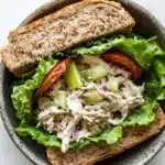 Loaded chicken salad sandwich - the image is a close-up of a sandwich in a gray bowl. the sandwich is made with two slices of toasted bread and is filled with a salad. the salad is made up of lettuce, diced tomatoes, celery, and other vegetables. the lettuce is a light green color and appears to be fresh and wilted. the tomatoes are red and appear to be seasoned with herbs and spices. the chicken is a creamy white color and is mixed in with the lettuce and tomatoes. the bowl is sitting on a white marble countertop.