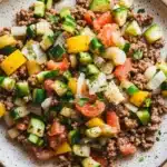 Low carb ground beef for dinner - the image is a close-up of a plate of food. the plate is white with a speckled pattern and is placed on a wooden table. the food appears to be a salad with a variety of ingredients, including ground beef, diced tomatoes, zucchini, yellow bell peppers, and chopped onions. the colors of the ingredients are vibrant and the dish looks fresh and appetizing.