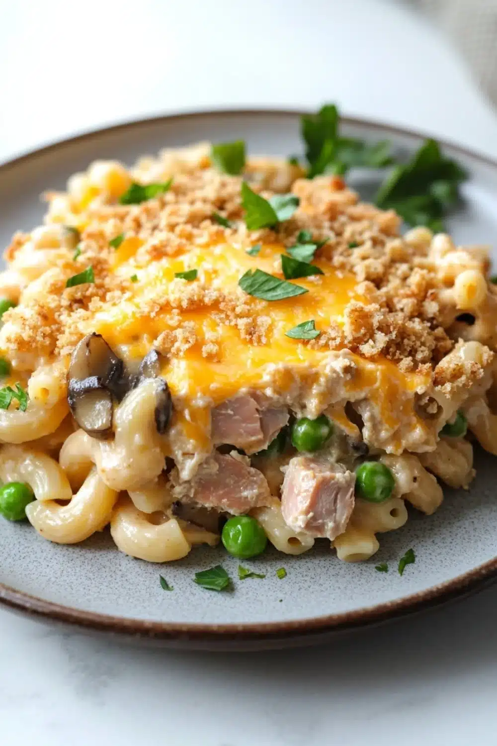 Lowfat tuna casserole - the image shows a plate of macaroni and cheese dish. the dish is served on a gray plate with a white tablecloth in the background. the macaronis are cooked al dente and are topped with a layer of melted cheese, sliced mushrooms, and chunks of ham. the cheese is crumbled and golden brown, and there are a few sprigs of parsley scattered around the plate. the overall appearance of the dish is savory and appetizing.