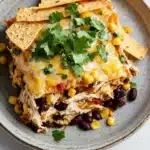 Make ahead chicken taco casserole - the image shows a plate of food with a layer of chicken enchilada on top. the dish is made up of shredded chicken, black beans, corn kernels, and cilantro. the chicken is cooked to a golden brown color and appears to be seasoned with herbs and spices. the tortilla chips are scattered on top of the dish, and there are a few sprigs of cilantro scattered around the plate. the plate is sitting on a white marble countertop.