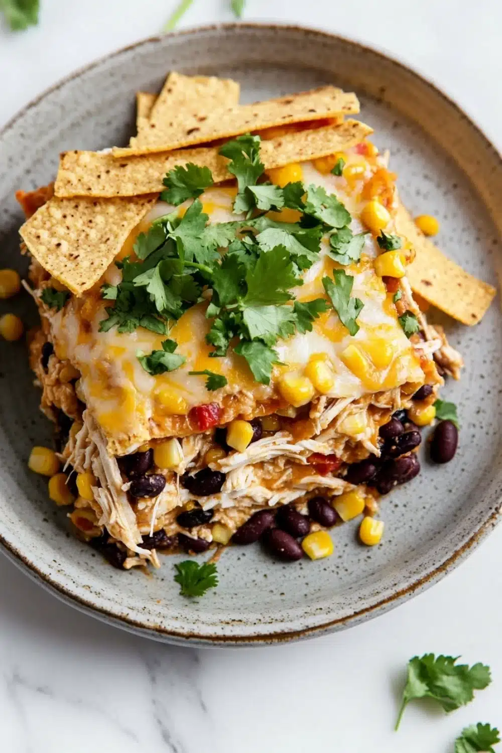 Make ahead chicken taco casserole - the image shows a plate of food with a layer of chicken enchilada on top. the dish is made up of shredded chicken, black beans, corn kernels, and cilantro. the chicken is cooked to a golden brown color and appears to be seasoned with herbs and spices. the tortilla chips are scattered on top of the dish, and there are a few sprigs of cilantro scattered around the plate. the plate is sitting on a white marble countertop.