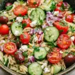 Mediterranean pasta salad with orzo - the image is a close-up of a bowl of pasta salad. the bowl is made of ceramic and has a green and brown speckled pattern. the pasta salad is made up of various types of pasta, including fusilli, cherry tomatoes, cucumbers, red onions, and feta cheese. the tomatoes are bright red and appear to be ripe and juicy. the cucumbers are sliced and arranged in a circular pattern, with some of them overlapping each other. the red onions are scattered throughout the salad, adding a pop of color and texture. the feta is crumbled and adds a creamy texture to the dish. the dish is garnished with fresh herbs, likely parsley or cilantro.
