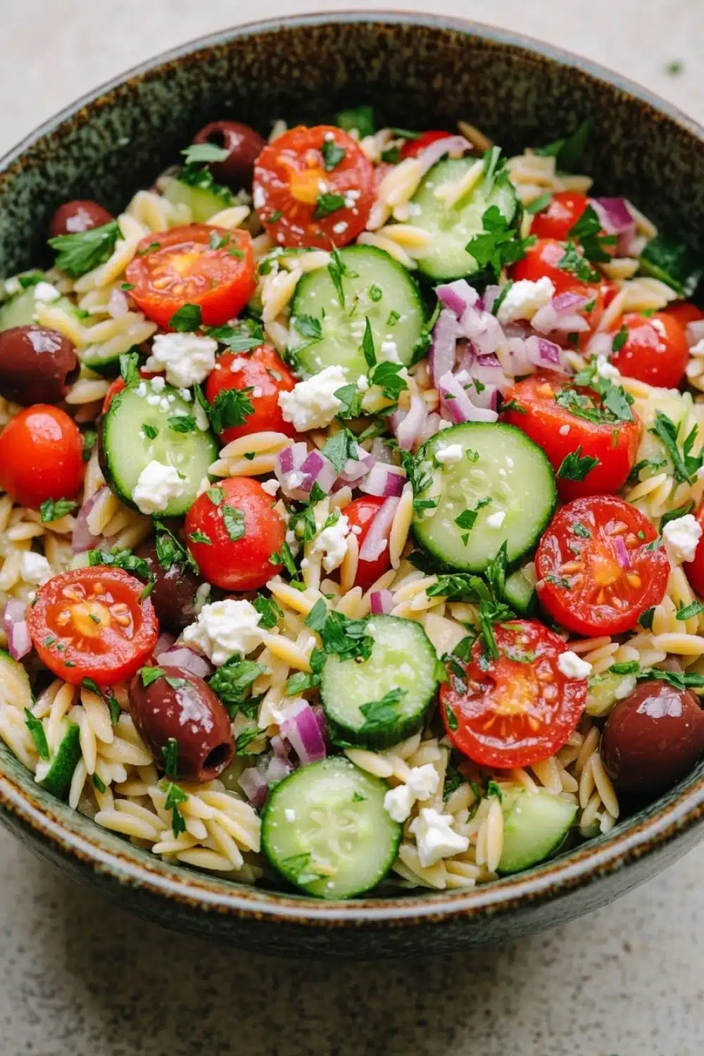 Mediterranean pasta salad with orzo - the image is a close-up of a bowl of pasta salad. the bowl is made of ceramic and has a green and brown speckled pattern. the pasta salad is made up of various types of pasta, including fusilli, cherry tomatoes, cucumbers, red onions, and feta cheese. the tomatoes are bright red and appear to be ripe and juicy. the cucumbers are sliced and arranged in a circular pattern, with some of them overlapping each other. the red onions are scattered throughout the salad, adding a pop of color and texture. the feta is crumbled and adds a creamy texture to the dish. the dish is garnished with fresh herbs, likely parsley or cilantro.