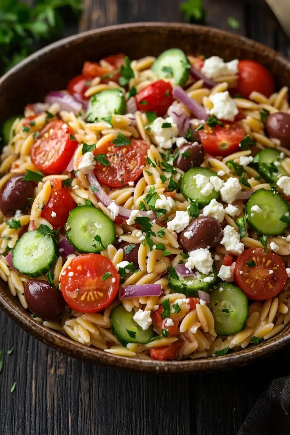 Mediterranean pasta salad with orzo - the image is a close-up of a bowl of pasta salad. the bowl is made of dark brown ceramic and is placed on a wooden table. the pasta salad is made up of fusilli, cherry tomatoes, cucumbers, red onions, and feta cheese. the tomatoes are bright red and the cucumbers are sliced into thin strips. the red onions are scattered throughout the salad, adding a pop of color and texture. the feta is crumbled and adds a creamy texture to the dish. there are also a few sprigs of parsley scattered around the bowl. the background is blurred, but it appears to be a rustic kitchen countertop.