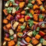 Mexican roasted root vegetables - the image shows a baking tray filled with roasted sweet potatoes and red onions. the sweet potatoes are cut into small cubes and are arranged in a single layer on the tray. the onions are sliced red and appear to be seasoned with herbs and spices. the tray is lined with parchment paper and is placed on a wooden table. the overall appearance of the dish is colorful and appetizing.