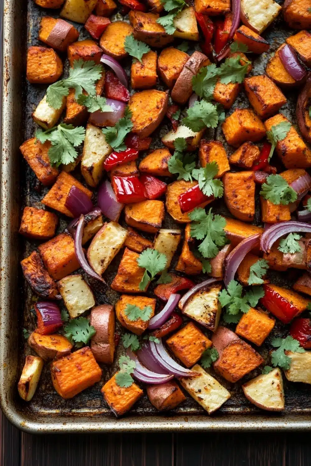 Mexican roasted root vegetables - the image shows a baking tray filled with roasted sweet potatoes and red onions. the sweet potatoes are cut into small cubes and are arranged in a single layer on the tray. the onions are sliced red and appear to be seasoned with herbs and spices. the tray is lined with parchment paper and is placed on a wooden table. the overall appearance of the dish is colorful and appetizing.