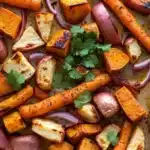 Mexican roasted root vegetables - the image shows a baking tray filled with roasted vegetables. the vegetables are cut into small cubes and are arranged in a single layer on the tray. the carrots are orange in color and appear to be seasoned with herbs and spices. there are also red onions and cilantro scattered throughout the tray, adding a pop of green color to the dish. the tray is lined with parchment paper and is sitting on a wooden surface.
