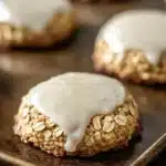Oatmeal cookie glaze - the image shows a close-up of a cookie on a baking tray. the cookie appears to be oatmeal raisin cookies with a golden brown crust and a drizzle of white icing on top. the icing is drizzled over the top of the cookie, creating a smooth and glossy texture. the tray is lined with parchment paper, and there are other cookies visible in the background. the overall mood of the image is warm and inviting.