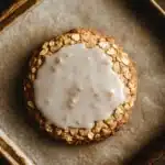 Oatmeal cookie glaze - the image shows a round cookie in a square baking tray. the cookie appears to be made with oats and nuts, and is covered in a creamy white icing. the tray is lined with parchment paper, and the background is a dark wooden surface. the overall mood of the image is warm and inviting.