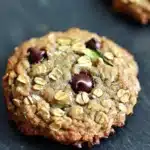 Oatmeal zucchini chocolate chip cookie - the image shows a close-up of a freshly baked oatmeal cookie on a black slate surface. the cookie is round and golden brown in color, with a crumbly texture. it is topped with chopped nuts and chocolate chips, giving it a crunchy appearance. in the background, there are more cookies visible, but they are slightly blurred. the overall mood of the image is warm and inviting.