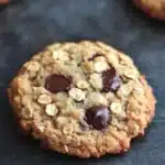 Oatmeal zucchini chocolate chip cookie - the image shows a close-up of a freshly baked oatmeal cookie on a dark grey surface. the cookie is round and golden brown in color, with a crumbly texture. it is topped with chopped nuts and chocolate chips, giving it a crunchy appearance. in the background, there are two more cookies visible, slightly blurred. the overall mood of the image is warm and inviting.