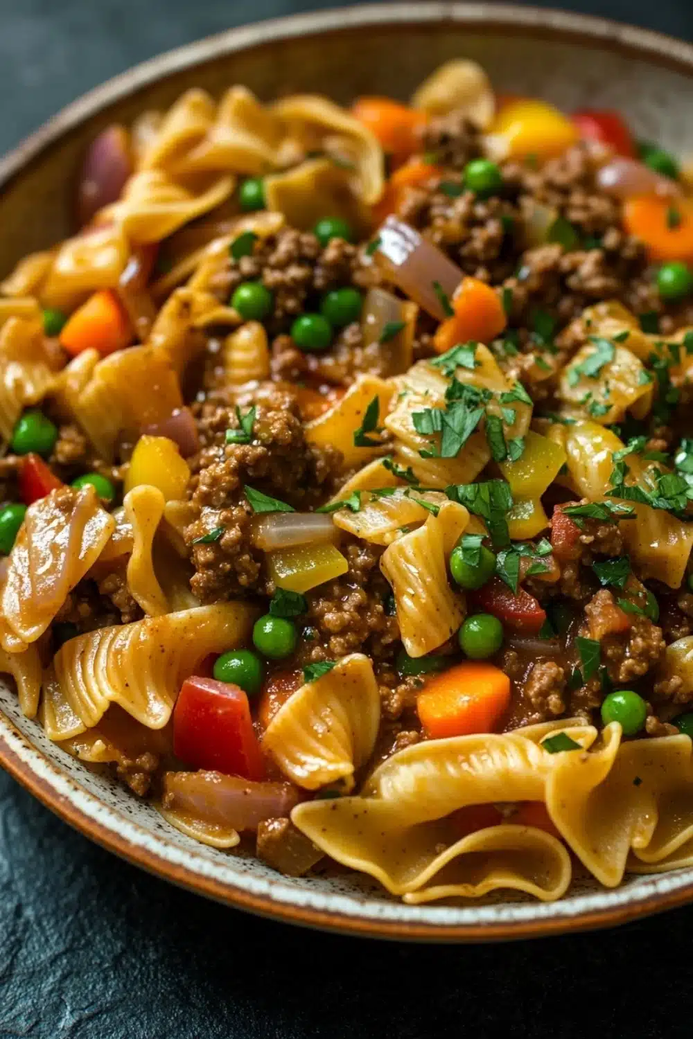 One pot beef noodle skillet - the image is a close-up of a bowl of pasta dish. the pasta appears to be fusilli, which is a type of spiral-shaped pasta, and is mixed with ground beef, peas, carrots, and onions. the dish is garnished with chopped parsley. the bowl is made of ceramic and is sitting on a dark blue tablecloth. the colors of the dish are vibrant and the texture looks fresh and appetizing.
