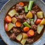One pot beef potatoes - the image is a close-up of a bowl of beef stew. the bowl is made of ceramic and has a speckled pattern on the rim. the stew is a rich brown color and appears to be made with chunks of beef, carrots, potatoes, onions, and celery. the beef is cooked medium-rare and looks tender and juicy. the vegetables are cut into small cubes and are arranged in a colorful and appetizing manner. the broth is thick and thick, and the dish looks thick and flavorful. the background is dark, making the colors of the stew stand out even more.
