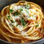 One pot carmelized onion pasta - the image shows a close-up of a bowl of spaghetti. the spaghetti is cooked al dente and is covered in a tomato sauce. the sauce is a deep red color and appears to be thick and creamy. on top of the spaghetti, there is a generous amount of grated parmesan cheese, which is sprinkled with chopped parsley. the bowl is made of dark-colored ceramic and is sitting on a dark wooden table. the background is blurred, but it seems to be a kitchen countertop.