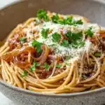 One pot carmelized onion pasta - the image is a close-up of a plate of spaghetti. the spaghetti is cooked al dente and is topped with a generous amount of tomato sauce and chunks of onions. the sauce is a deep red color and appears to be thick and creamy. the dish is garnished with fresh parsley and grated parmesan cheese. the plate is made of a light-colored ceramic bowl with a speckled pattern. the background is a white countertop with a blue and white striped napkin.