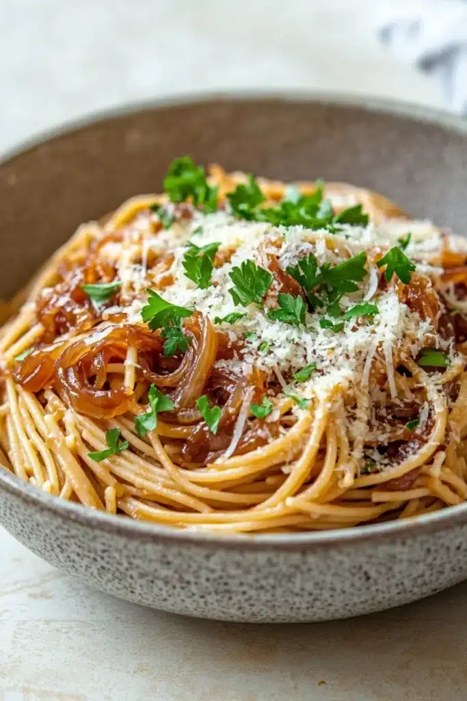 One pot carmelized onion pasta - the image is a close-up of a plate of spaghetti. the spaghetti is cooked al dente and is topped with a generous amount of tomato sauce and chunks of onions. the sauce is a deep red color and appears to be thick and creamy. the dish is garnished with fresh parsley and grated parmesan cheese. the plate is made of a light-colored ceramic bowl with a speckled pattern. the background is a white countertop with a blue and white striped napkin.