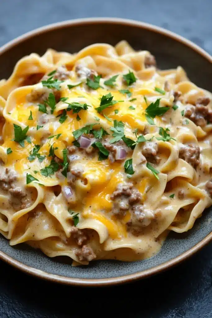 One pot creamy beef and noodles - the image shows a plate of tagliatelle pasta dish. the pasta is cooked al dente and is topped with a creamy sauce and chunks of ground beef. the sauce is a light yellow color and appears to be thick and creamy. the dish is garnished with a sprig of fresh parsley. the plate is brown and is sitting on a dark grey countertop.
