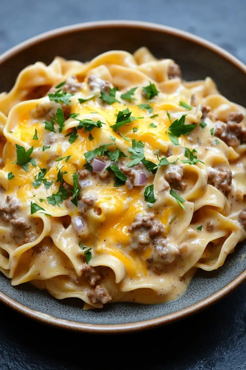One pot creamy beef and noodles - the image shows a plate of tagliatelle pasta dish. the pasta is cooked al dente and is topped with a creamy sauce and chunks of ground beef. the sauce is a light yellow color and appears to be thick and creamy. the dish is garnished with a sprig of fresh parsley. the plate is brown and is sitting on a dark grey countertop.