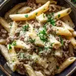 One pot creamy garlic butter beef pasta - the image is a close-up of a bowl of pasta dish. the pasta appears to be penne, which is a type of penne pasta, and it is cooked al dente. the dish is garnished with chopped parsley and grated parmesan cheese. the bowl is made of dark blue ceramic and is sitting on a dark wooden surface. there are a few sprigs of parsley scattered around the bowl, adding a pop of green color to the dish.