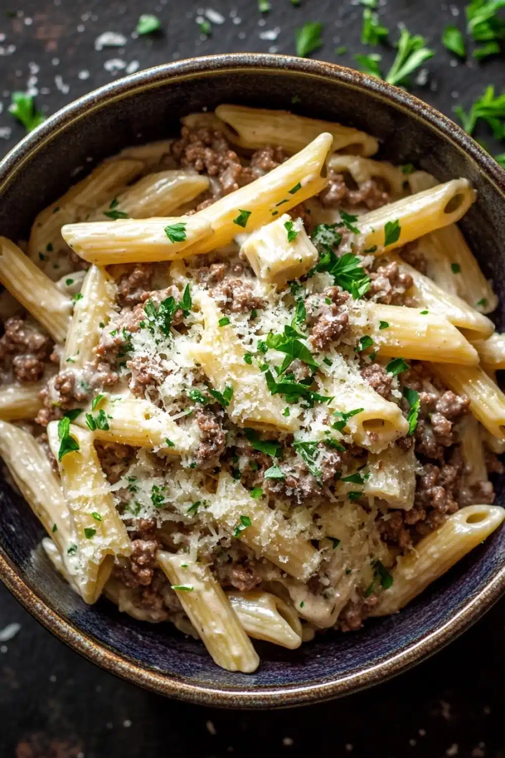 One pot creamy garlic butter beef pasta - the image is a close-up of a bowl of pasta dish. the pasta appears to be penne, which is a type of penne pasta, and it is cooked al dente. the dish is garnished with chopped parsley and grated parmesan cheese. the bowl is made of dark blue ceramic and is sitting on a dark wooden surface. there are a few sprigs of parsley scattered around the bowl, adding a pop of green color to the dish.