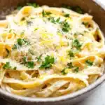 One pot creamy lemon pasta - the image is a close-up of a bowl of tagliatelle pasta. the pasta is cooked al dente and is topped with a generous amount of grated parmesan cheese. the dish is garnished with fresh parsley leaves. the bowl is made of ceramic and is sitting on a white marble countertop. the background is blurred, making the pasta the focal point of the image.