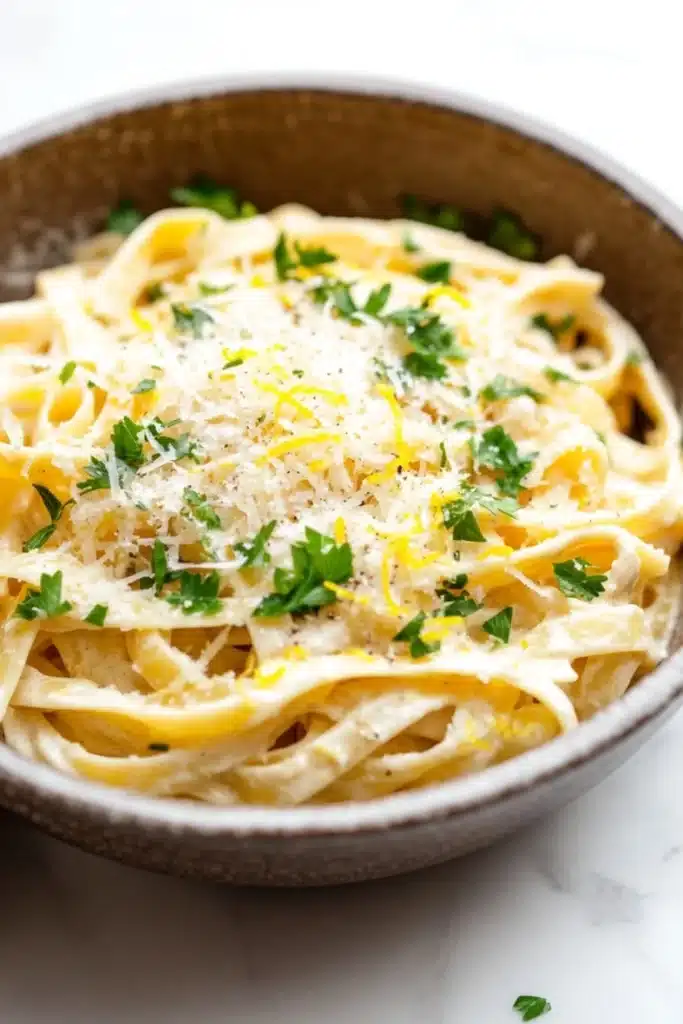 One pot creamy lemon pasta - the image is a close-up of a bowl of tagliatelle pasta. the pasta is cooked al dente and is topped with a generous amount of grated parmesan cheese. the dish is garnished with fresh parsley leaves. the bowl is made of ceramic and is sitting on a white marble countertop. the background is blurred, making the pasta the focal point of the image.