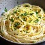 One pot creamy lemon pasta - the image is a close-up of a bowl of spaghetti. the spaghetti is cooked al dente and is covered in a creamy sauce. the sauce appears to be a light yellow color and is drizzled over the top of the spaghetti. there are small pieces of lemon zest scattered throughout the dish, adding a pop of green color to the dish. the bowl is made of ceramic and has a speckled pattern on the sides. the background is dark, making the spaghetti stand out.