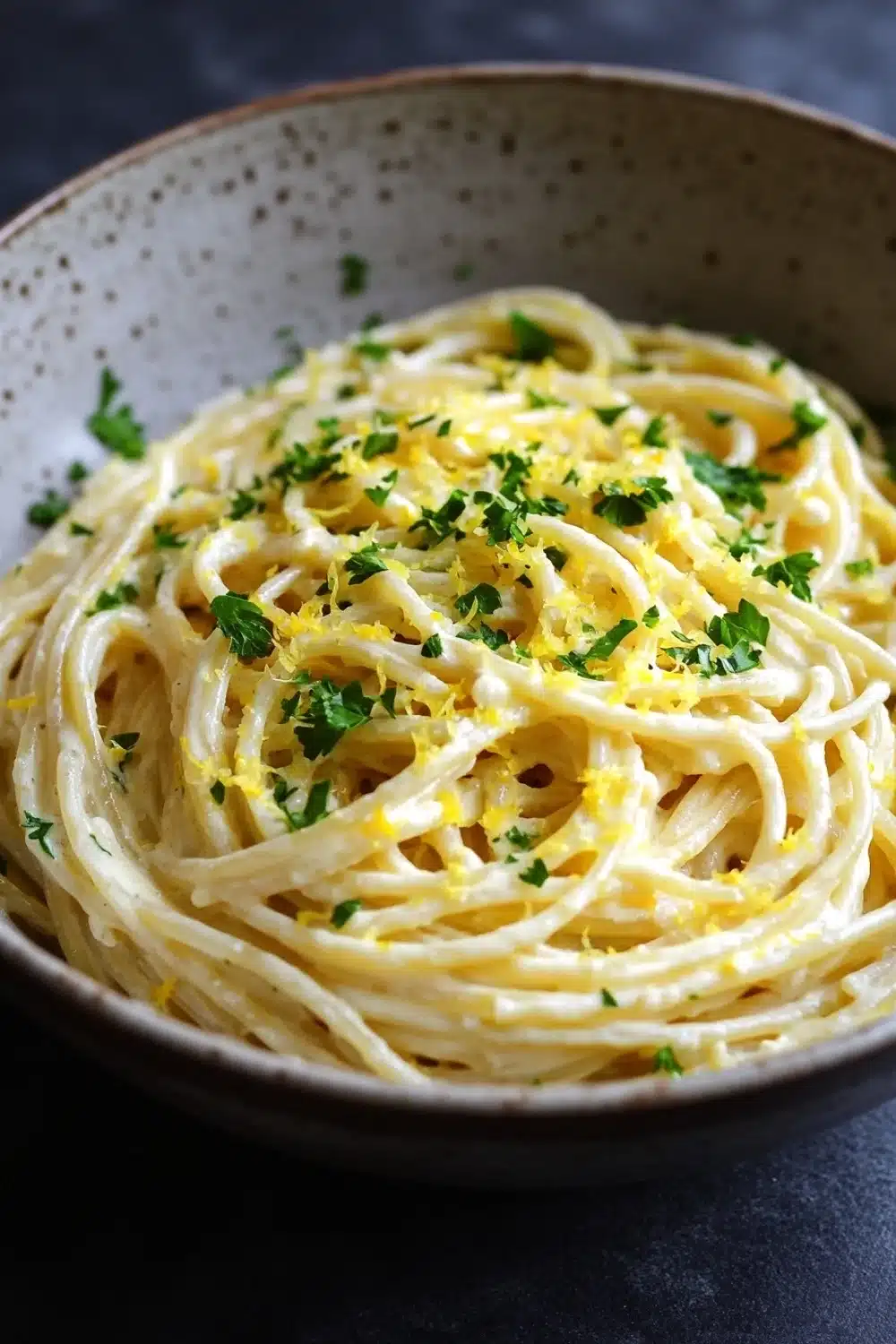 One pot creamy lemon pasta - the image is a close-up of a bowl of spaghetti. the spaghetti is cooked al dente and is covered in a creamy sauce. the sauce appears to be a light yellow color and is drizzled over the top of the spaghetti. there are small pieces of lemon zest scattered throughout the dish, adding a pop of green color to the dish. the bowl is made of ceramic and has a speckled pattern on the sides. the background is dark, making the spaghetti stand out.