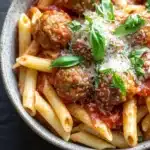 One pot pasta and meatballs - the image is a close-up of a bowl of penne pasta with meatballs and tomato sauce. the pasta is cooked al dente and is topped with a generous amount of tomato sauce and sprinkled with grated parmesan cheese. the meatballs are golden brown and appear to be seasoned with herbs and spices. the dish is garnished with fresh basil leaves. the bowl is sitting on a dark grey surface.