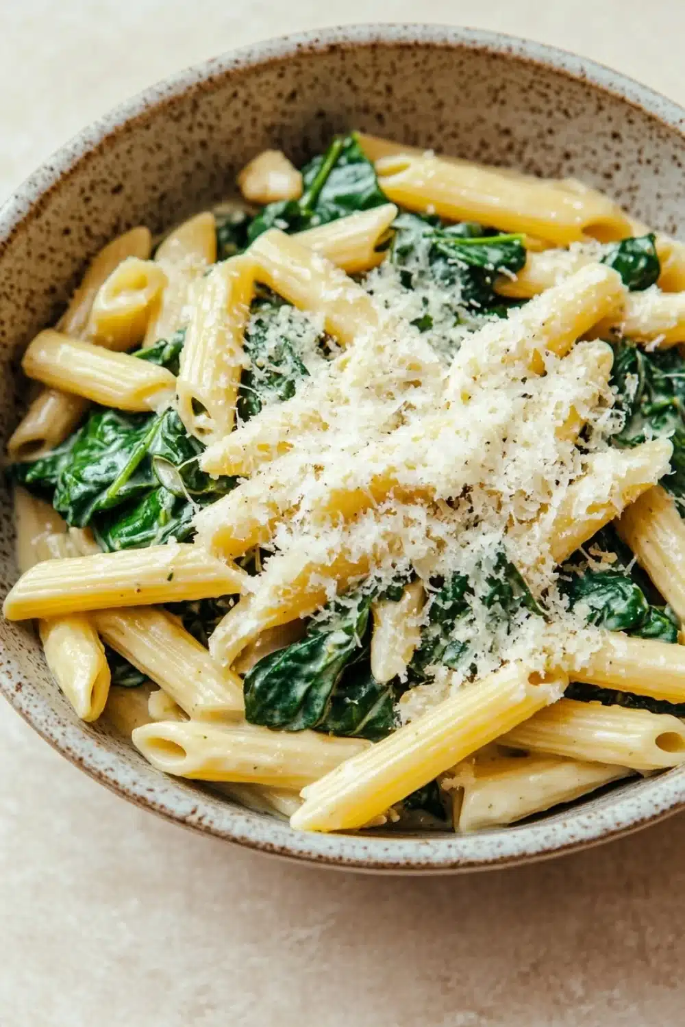 One pot pasta spinach - the image is a close-up of a bowl of pasta dish. the pasta appears to be penne or penne, and it is cooked al dente. the dish is garnished with fresh spinach leaves and grated parmesan cheese. the bowl is made of ceramic and has a light brown color. the background is a beige-colored tablecloth.