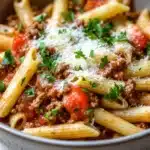 One pot pasta with meat sauce - the image is a close-up of a bowl of pasta dish. the pasta appears to be penne, which is a type of penne pasta. the dish is made with ground beef, tomato sauce, and chunks of tomato. the meat is cooked in a tomato-based sauce and is topped with grated parmesan cheese. there are also chunks of red bell peppers scattered throughout the dish, adding a pop of color. the bowl is white with a speckled pattern and is sitting on a wooden table.