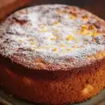 Orange french yogurt cake - the image is a close-up of a round cake on a green plate. the cake appears to be a chocolate cake with a golden brown crust and a layer of orange zest on top. the top of the cake is dusted with a light dusting of powdered sugar. the plate is sitting on a wooden table with a wooden cutting board in the background. the overall mood of the image is warm and inviting.