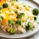Oven baked chicken broccoli rice casserole - the image shows a plate of food with a serving of rice and broccoli. the rice is white and fluffy, and the broccoli is bright green. on top of the rice, there is a layer of melted cheese that is golden brown and bubbly. the cheese appears to be melted and has a crumbly texture. the plate is light brown with a speckled pattern around the edges. the background is a white marble countertop.