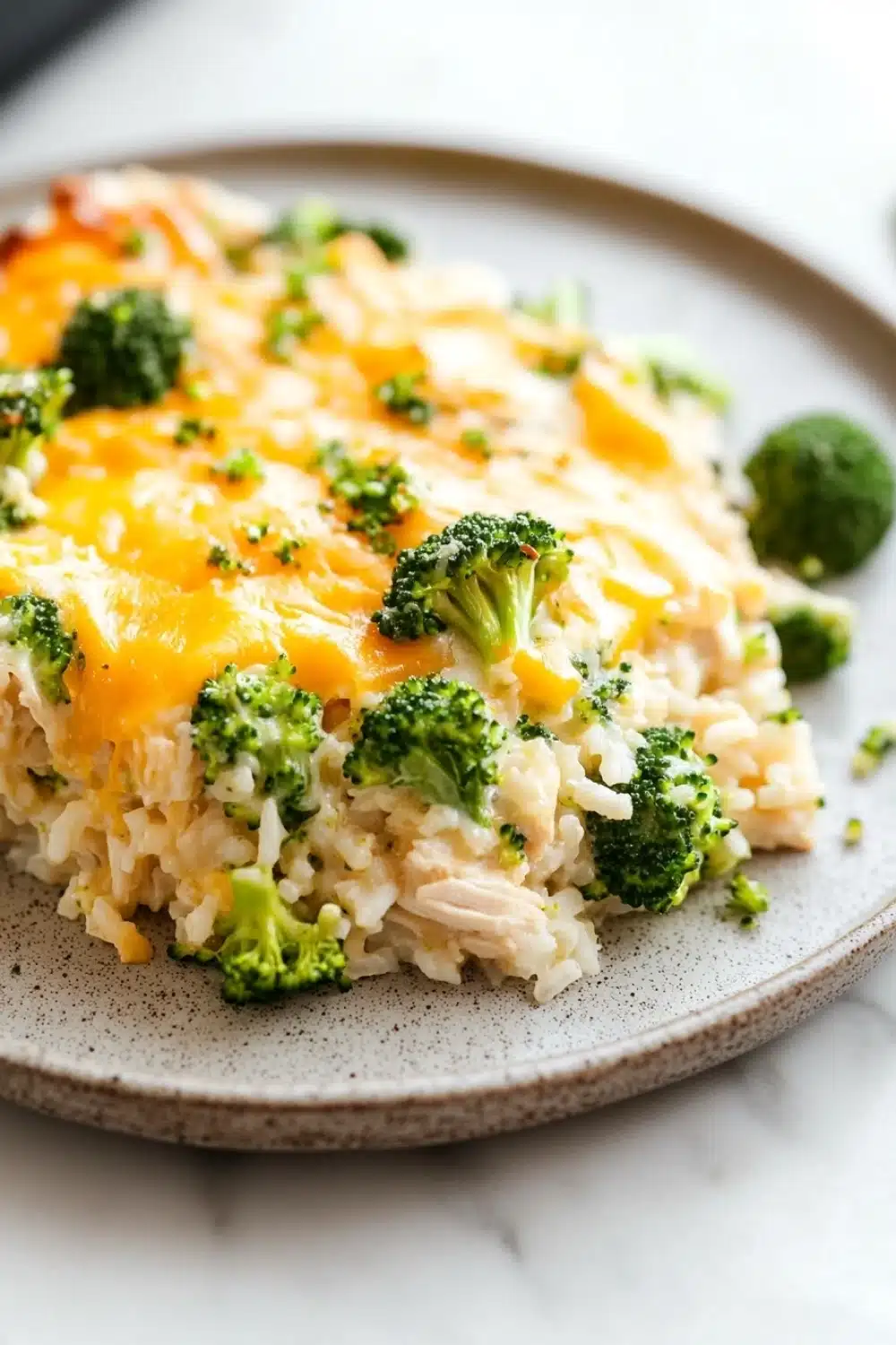 Oven baked chicken broccoli rice casserole - the image shows a plate of food with a serving of rice and broccoli. the rice is white and fluffy, and the broccoli is bright green. on top of the rice, there is a layer of melted cheese that is golden brown and bubbly. the cheese appears to be melted and has a crumbly texture. the plate is light brown with a speckled pattern around the edges. the background is a white marble countertop.