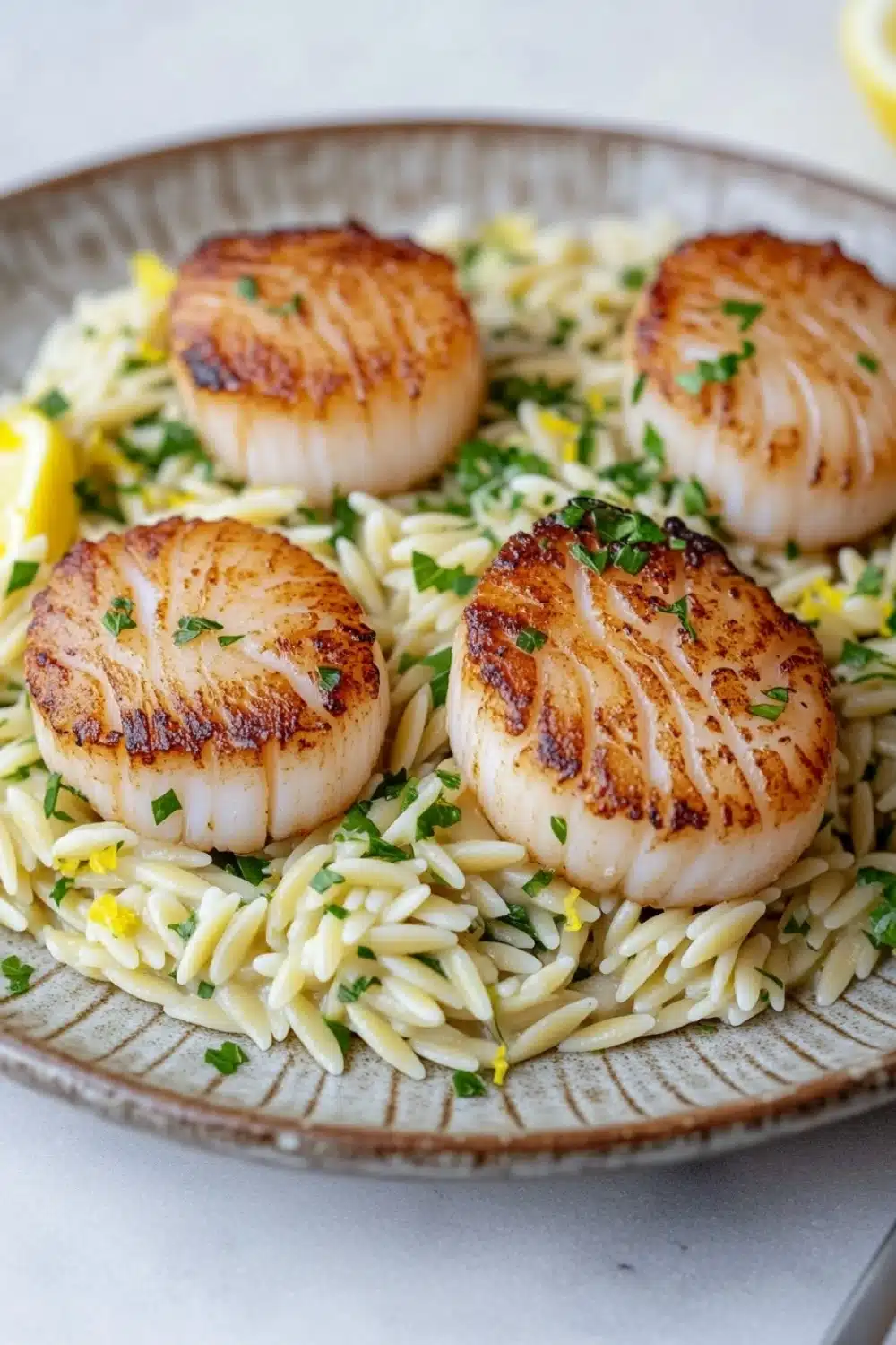 Pan seared scallops with orzo - the image shows a plate of cooked scallops on top of a bed of fusilli pasta. the scalls are golden brown and appear to be seasoned with herbs and spices. the pasta is a light yellow color and is scattered throughout the dish. there are also a few lemon wedges on the side of the plate. the plate is sitting on a white tablecloth and there is a fork resting on the right side.
