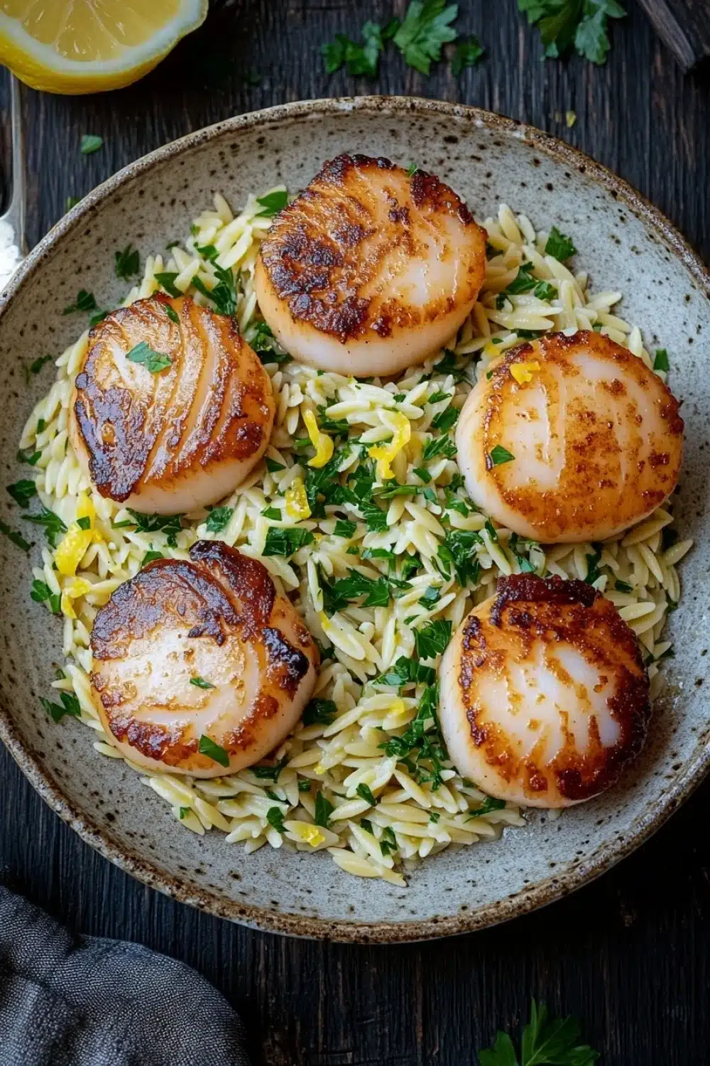 Pan seared scallops with orzo - the image shows a plate of cooked scallops on top of a bed of fusilli pasta. the plate is made of a light-colored ceramic bowl with a speckled pattern. the scallop shells are golden brown and appear to be seasoned with herbs and spices. the pasta is a mix of yellow and white, and there are small pieces of lemon scattered throughout the dish. there are also a few sprigs of parsley scattered around the plate, adding a pop of green color to the dish and a slice of lemon on the side. a fork and knife are placed on the wooden table next to the plate.
