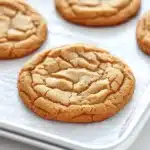 Peanut butter cookie no butter - the image shows a tray of freshly baked peanut butter cookies. the cookies are round and golden brown in color, with a criss-cross pattern on top. they are arranged in a single layer on a white parchment paper lined baking tray. the tray is placed on a marble countertop. the background is blurred, but it appears to be a kitchen countertop with other baking utensils visible.