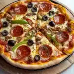 Pellet grill cast iron pizza - the image is of a large pepperoni pizza on a wooden cutting board. the pizza is round and has a golden brown crust. it is topped with sliced pepperoni, black olives, and green bell peppers. there are also small pieces of sausage scattered throughout the pizza. the cutting board is placed on a white marble countertop. in the background, there is a small bowl of pesto sauce.