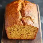 Pumpkin bread applesauce - the image shows a freshly baked loaf of bread on a wooden cutting board. the bread appears to be golden brown in color and has a crumbly texture. it is sitting on a dark grey countertop with a textured surface. the loaf is cut in half, revealing the soft, fluffy interior. the edges of the bread are slightly curled, indicating that it has been freshly baked.
