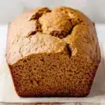 Pumpkin bread gf - the image shows a freshly baked loaf of bread on a wooden cutting board. the bread appears to be golden brown in color and has a slightly crumbly texture. it is sitting on a white marble countertop with a blurred background. the loaf is cut in half, revealing the soft, fluffy interior. the edges of the bread are slightly curled, indicating that it has been freshly baked.